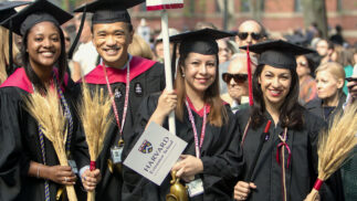 Four graduates of Harvard Extension School at commencement holding signs and two of the symbols of the School, wheat and a lamp.