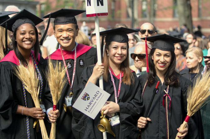 Four graduates of Harvard Extension School at commencement holding signs and two of the symbols of the School, wheat and a lamp.