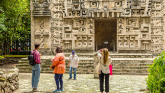 Four tourists at a Maya temple taking pictures and viewing the relief.