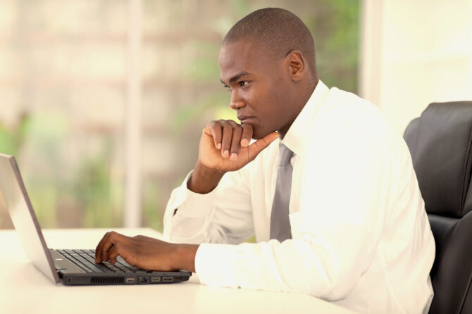 Young adult wearing a shirt and tie looking thoughtfully at a notebook computer.