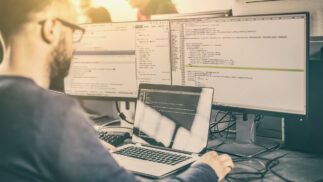 Computer scientist working at a notebook computer with large external monitors displaying code.