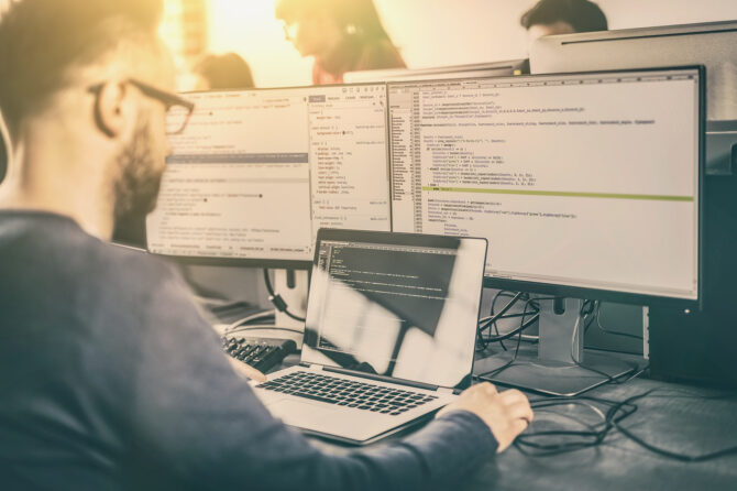 Computer scientist working at a notebook computer with large external monitors displaying code.