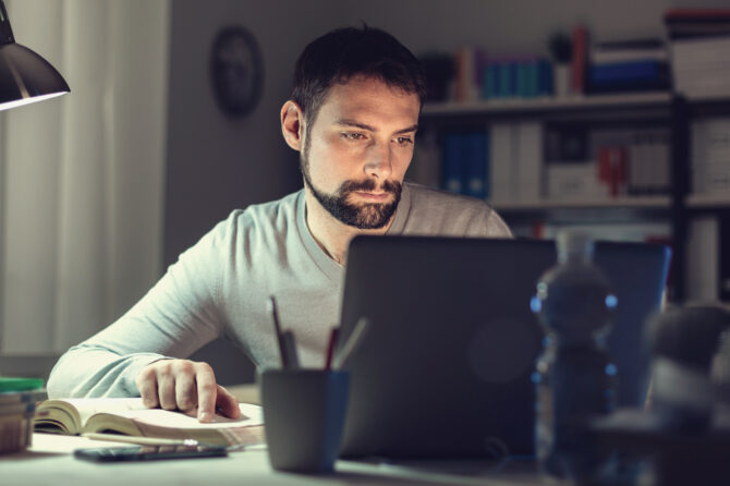 Person writing a paper on a notebook computer using a literary source as a reference.