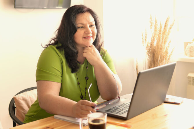 A woman sits in front of a laptop, looking thoughtful.
